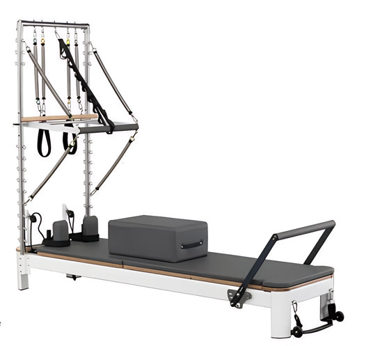 Pilates reformer machine with a white frame, dark gray padded bed and block, wooden accents, and a metal spring frame system on a plain white background.